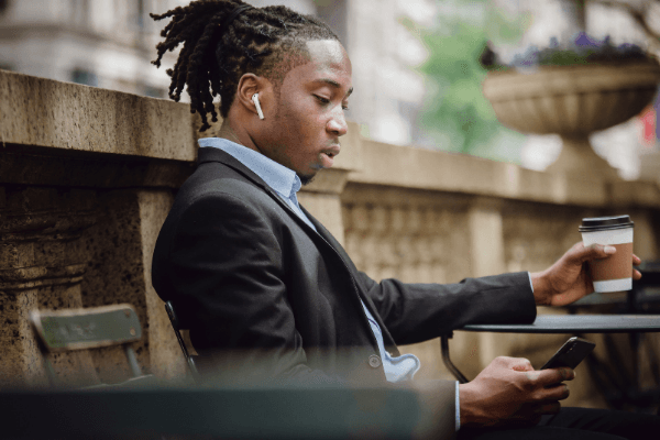 Young man in a suit with headphones on looking at his phone and drinking coffee outdoors