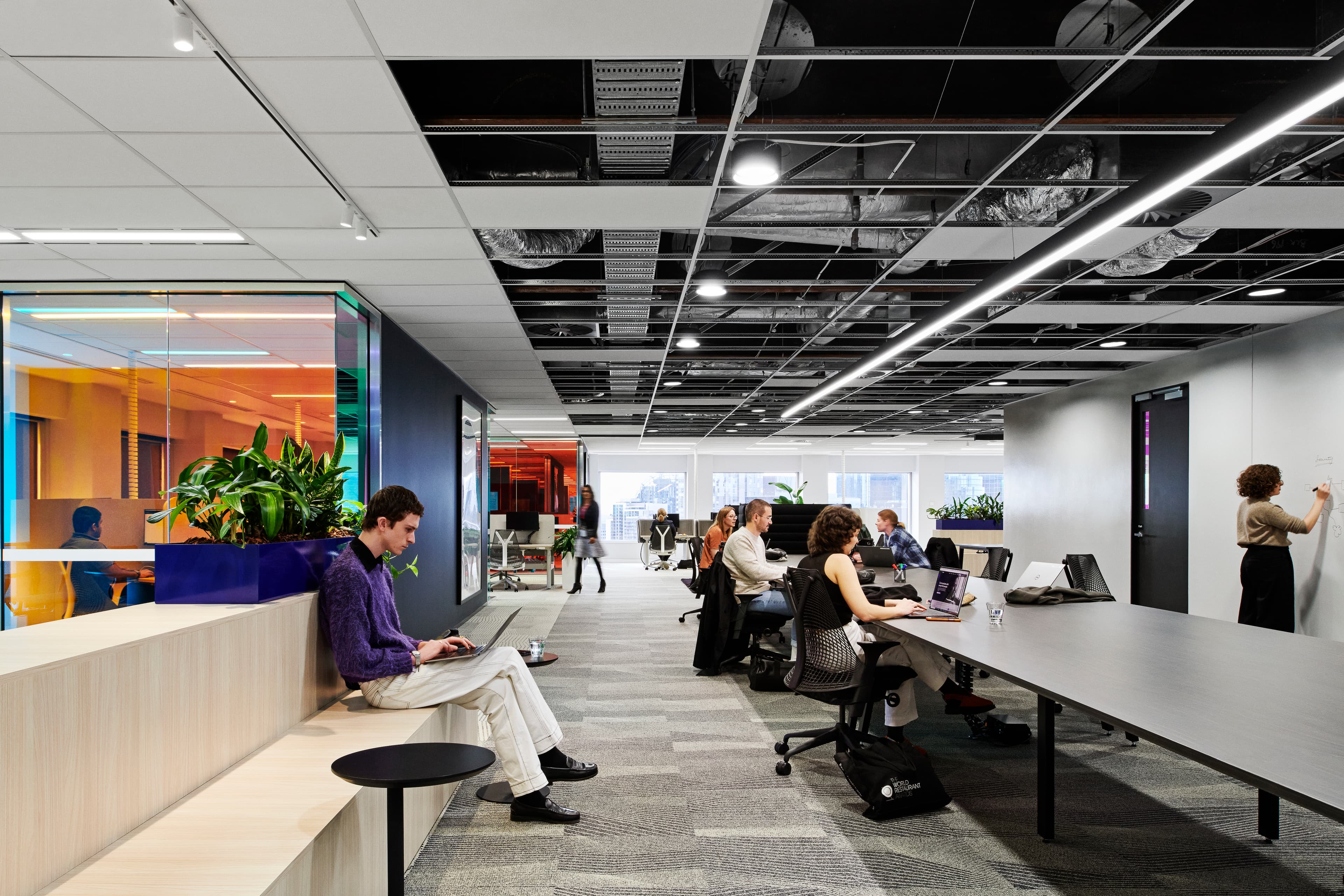 Open workplace with a man sitting on a bench working, a group of people working on a long open table, and a woman writing on a wall whiteboard